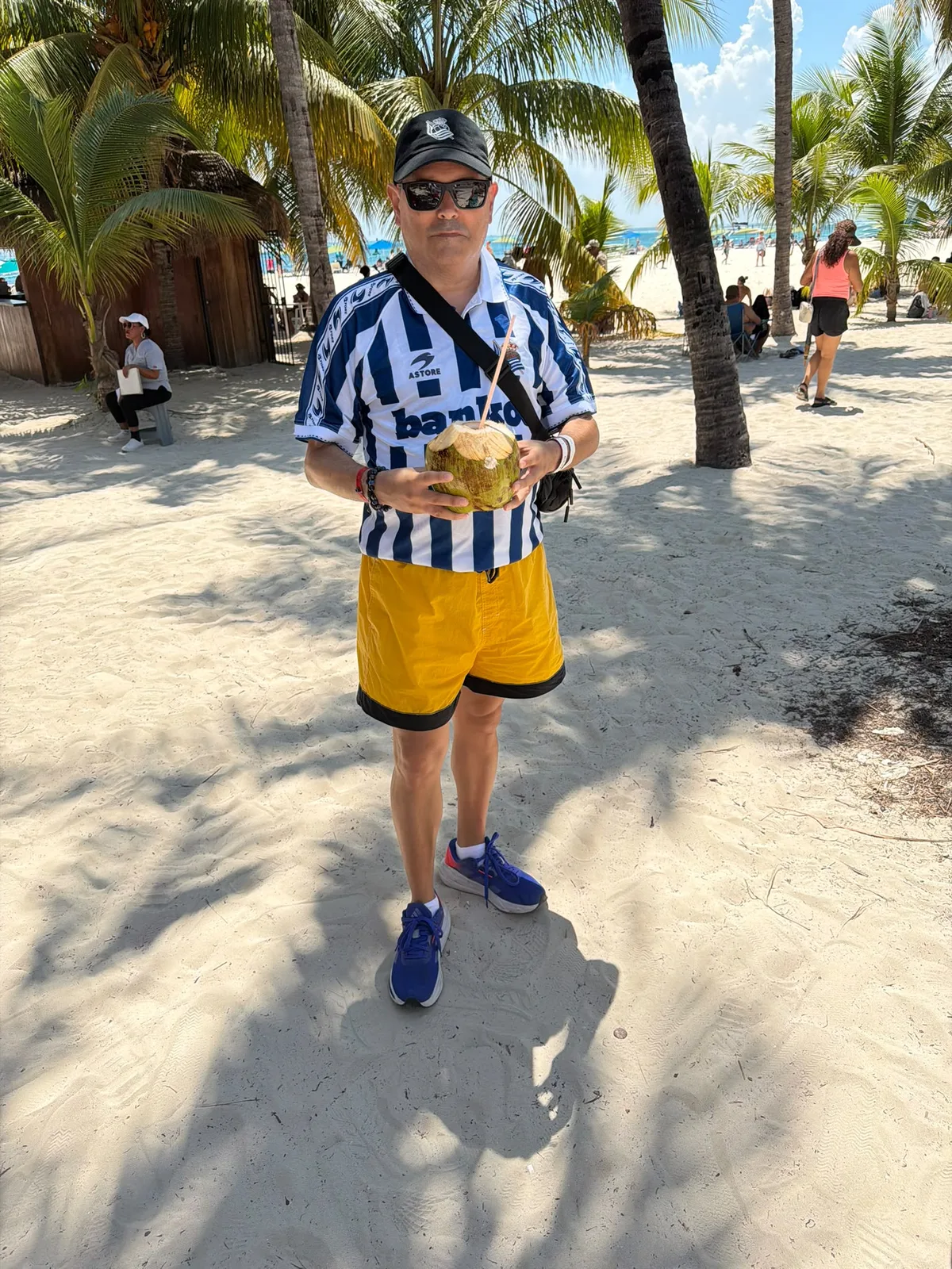 Socio de la peña con gorra y camiseta de la Real Sociedad sujetando un coco en una playa de Cancún, entre palmeras