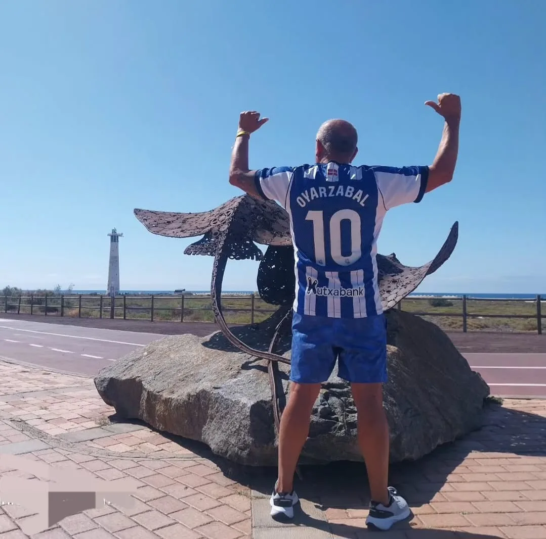 Socio de la peña con la camiseta de Oyarzabal y la Real Sociedad, ante una escultura y un faro en Fuerteventura