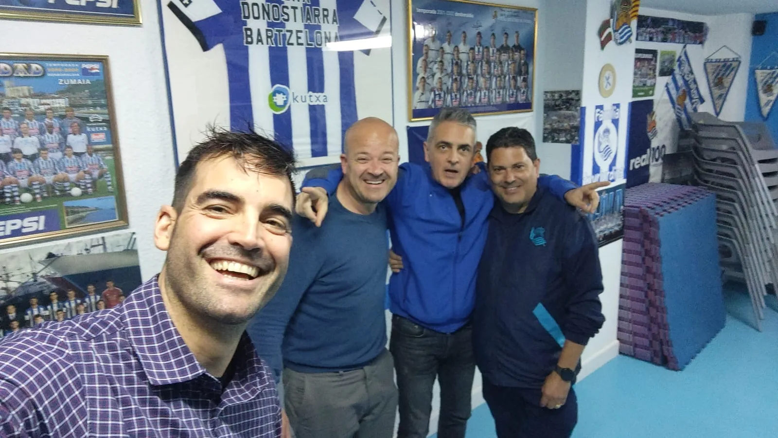 Socios de la peña en su local junto a la bandera Peña Donostiarra Bartzelona y fotos de equipo enmarcadas