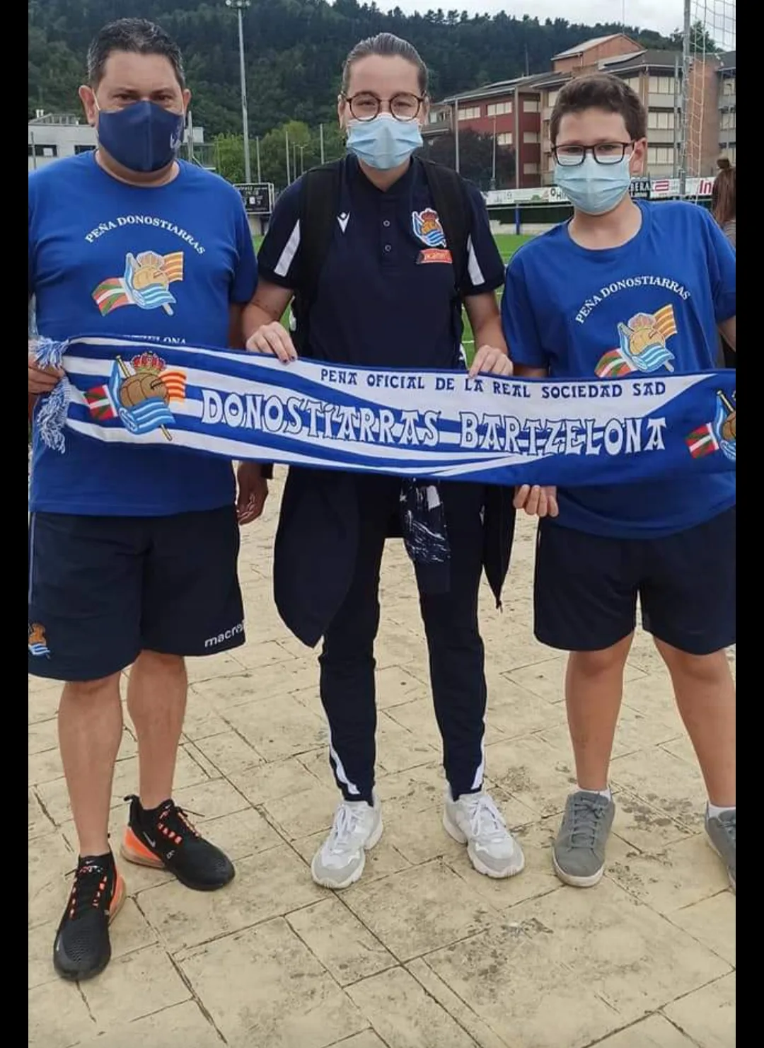 Socio de la peña con dos jóvenes socios sujetando la bufanda junto a un campo de fútbol
