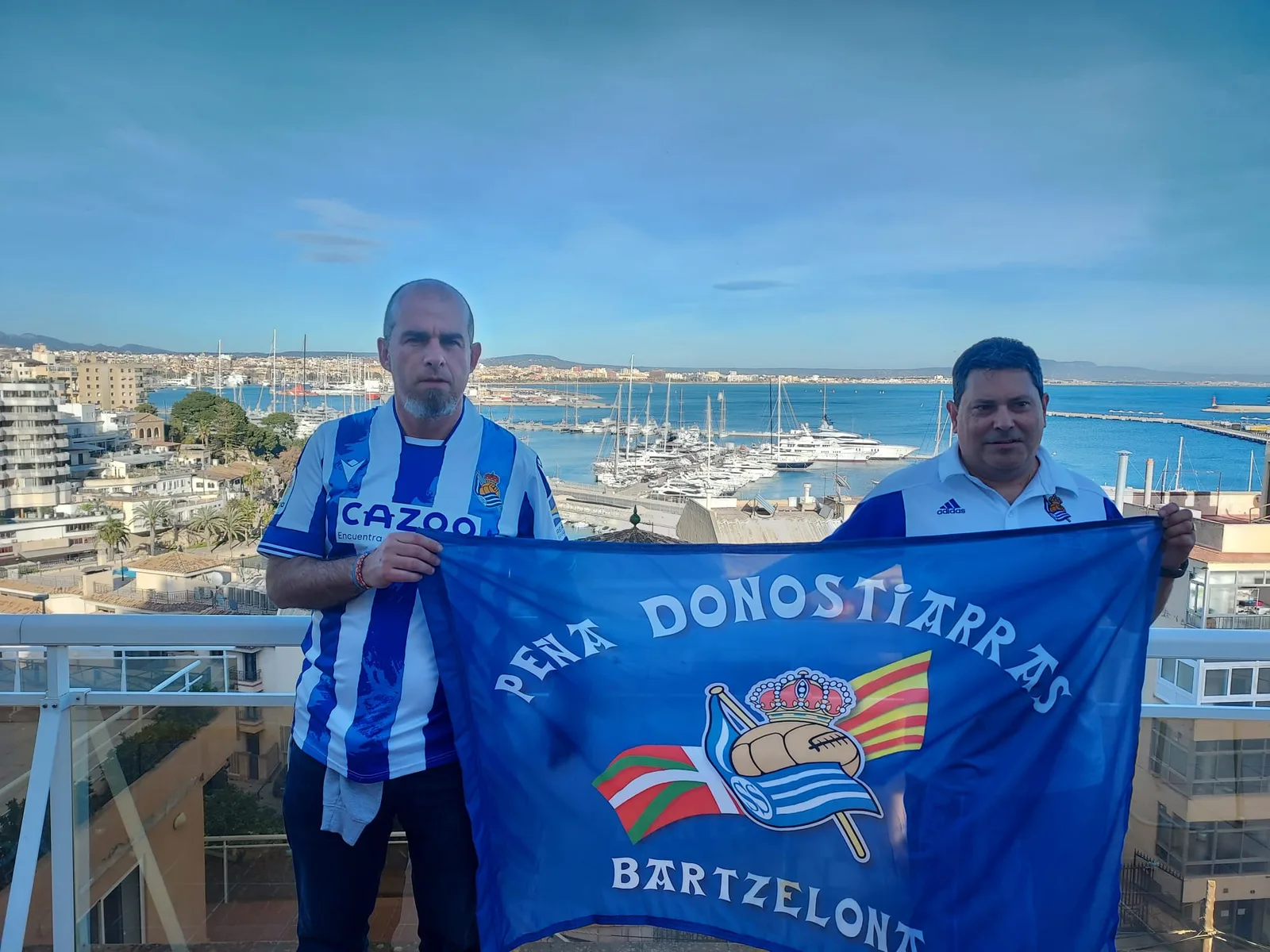 Socios de la peña con la bandera de la Peña Donostiarras Bartzelona y el puerto deportivo de Palma al fondo