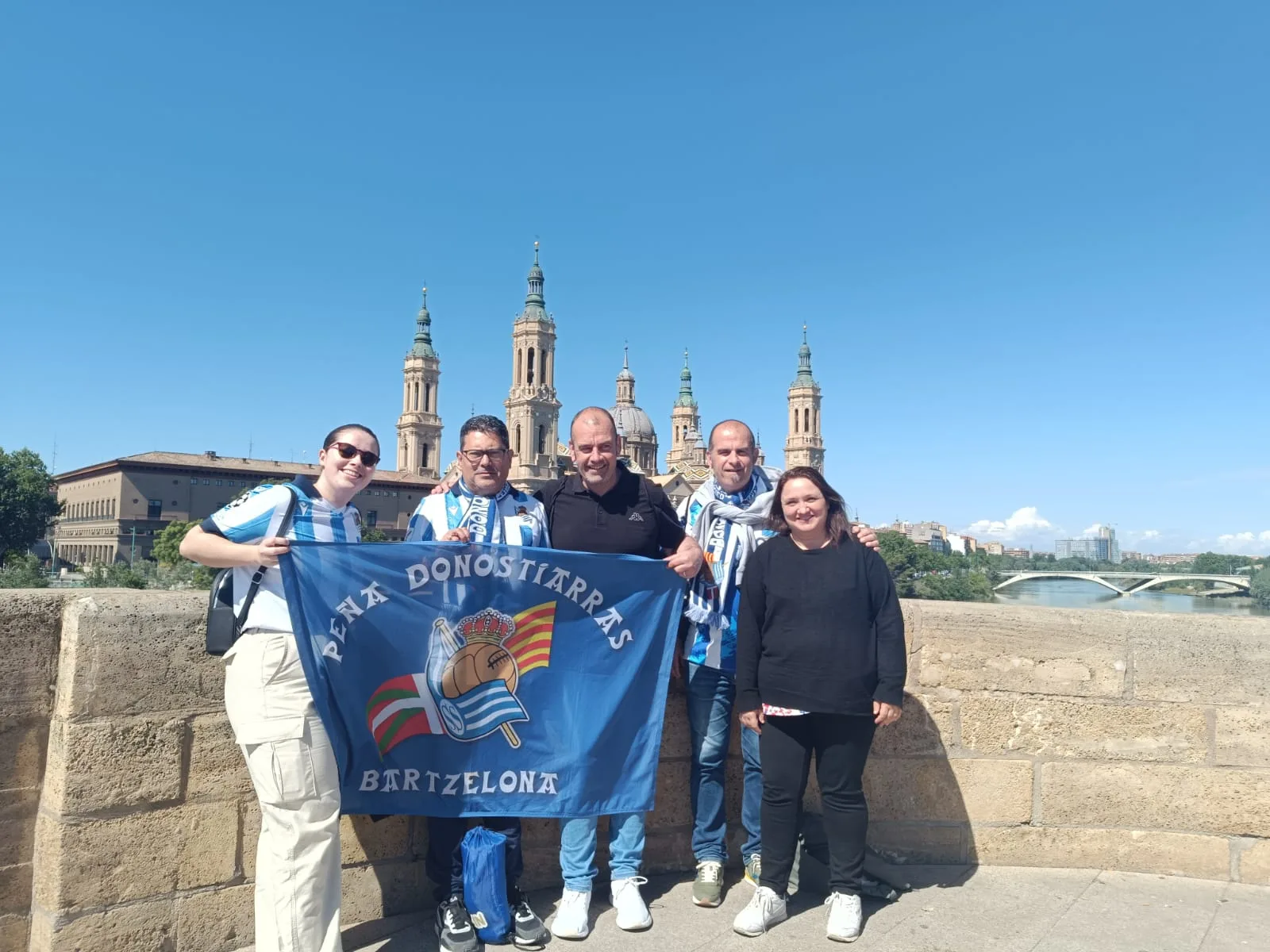 Socios con la bandera de la Peña Donostiarras Bartzelona en un puente sobre el Ebro, con la Basílica del Pilar al fondo
